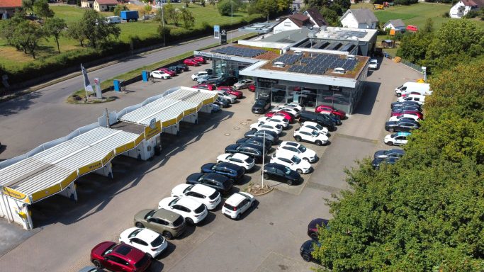 Parkplatz mit vielen Autos und einem Gebäuden inklusive der Waschanlage. Der Winkel entspricht einer Drohnenaufnahme.