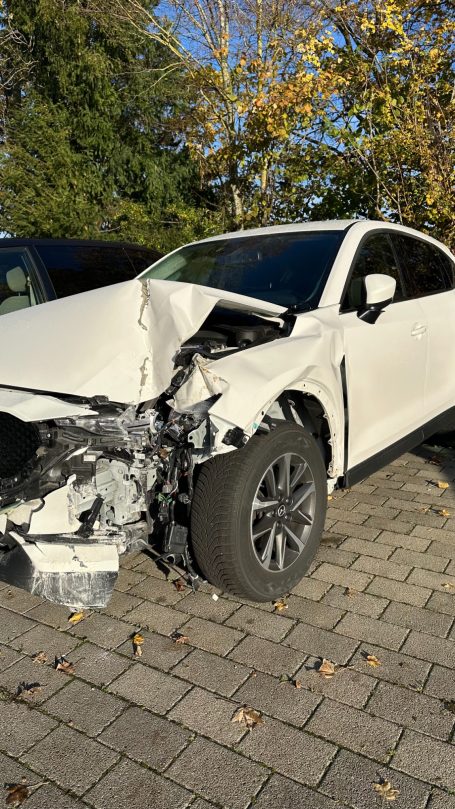 Ein beschädigter weißer SUV mit einem frontalen Unfallschaden steht auf einem Parkplatz.
